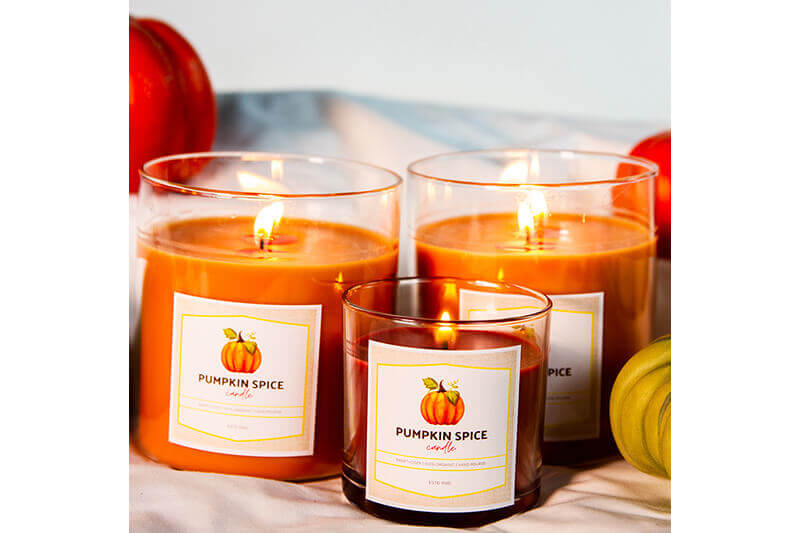 Three cylinder orange candles with custom-printed labels and pumpkins in the background.