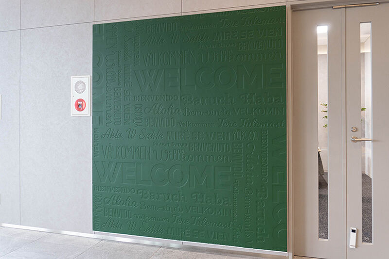 Wall with large square green embossed wall art with the word for "welcome" in many languages