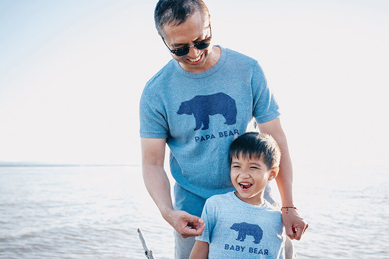 Father standing behind son on beach, each wearing light blue t-shirts with the words "Papa Bear" and "Baby Bear" on them.