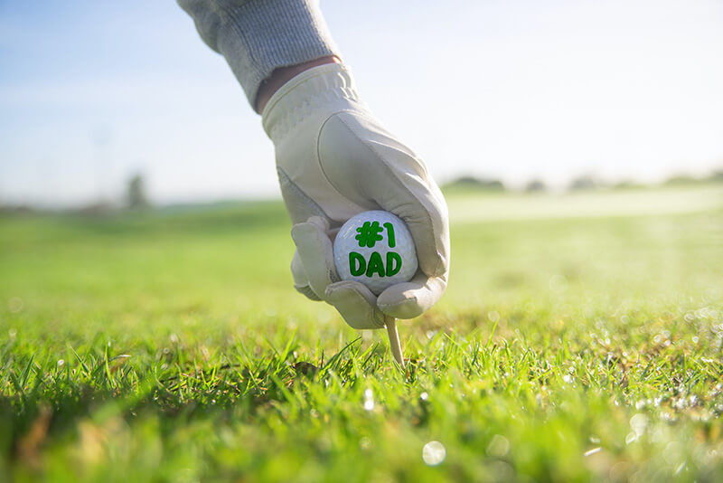 Mano con guante colocando una pelota de golf que dice "#1 Dad" ("papá N° 1") sobre el tee en el césped. 