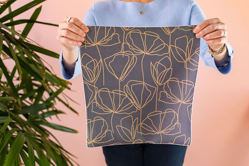 Torso of a woman holding a square pattern sample in front of her. The sample has a purple and yellow design.