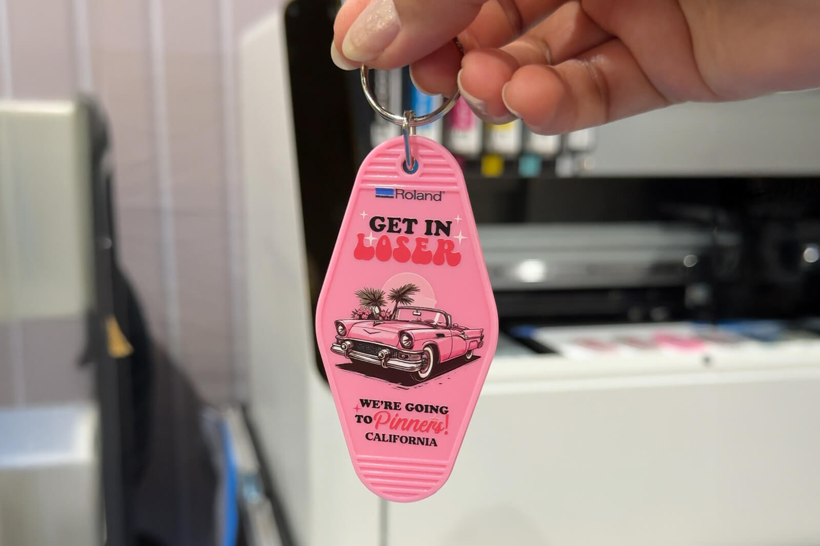 Hand holding single keychain with "Get in Loser" and car graphics in front of a Roland DG printer.