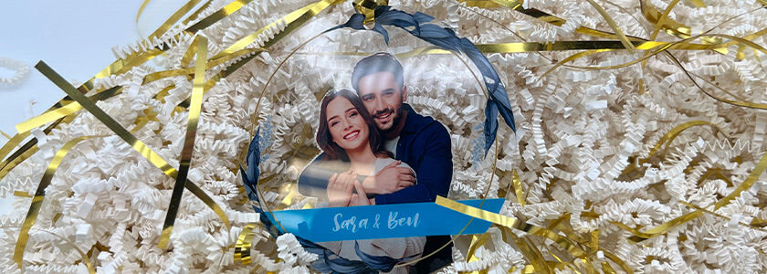 Ornamento de acrílico transparente con la imagen de una pareja y el texto “Sara y Ben”, sobre papel triturado.
