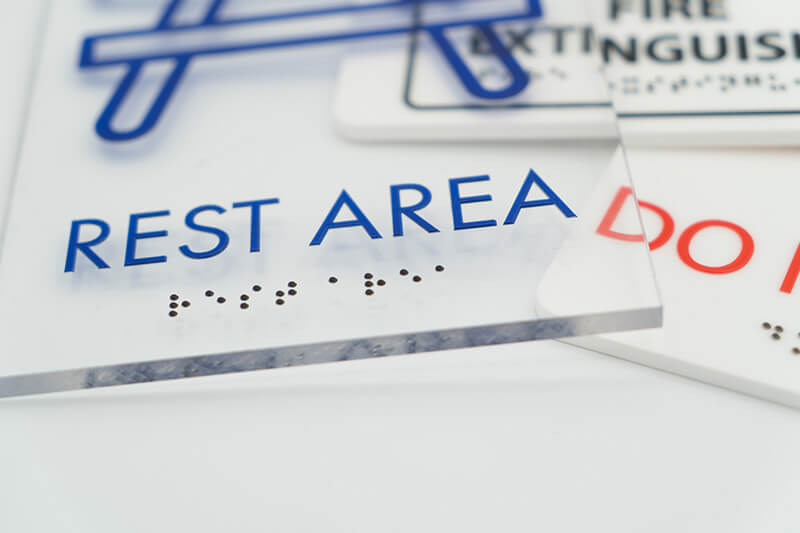 Primer plano de letreros de acrílico transparente con los mensajes "Área de Descanso", "No Entre" y "Extintor de Incendios", junto con su equivalente en braille.