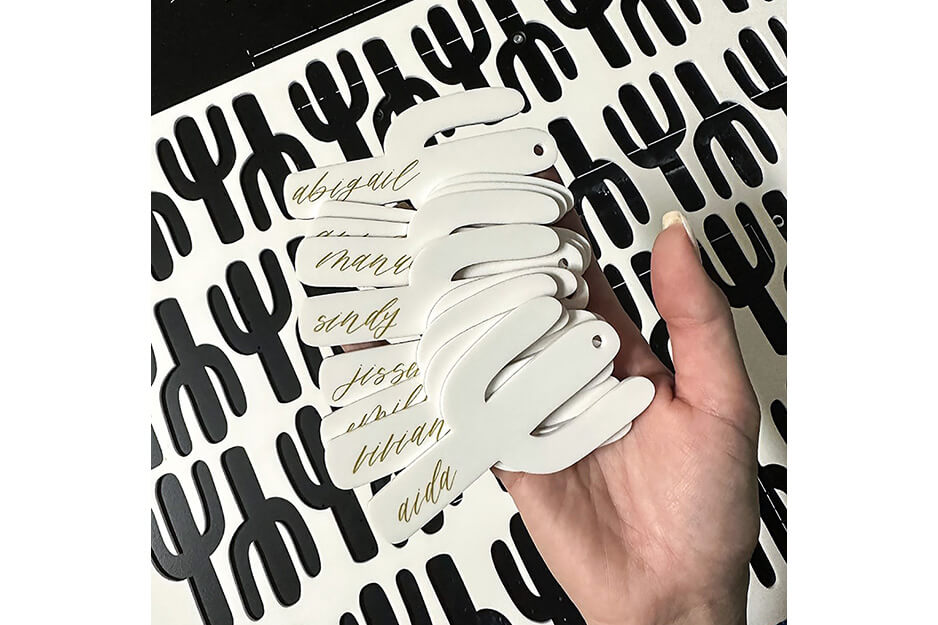 Hand holding white cactus cut outs with names printed on them against background of the white plastic the cactus were removed from.
