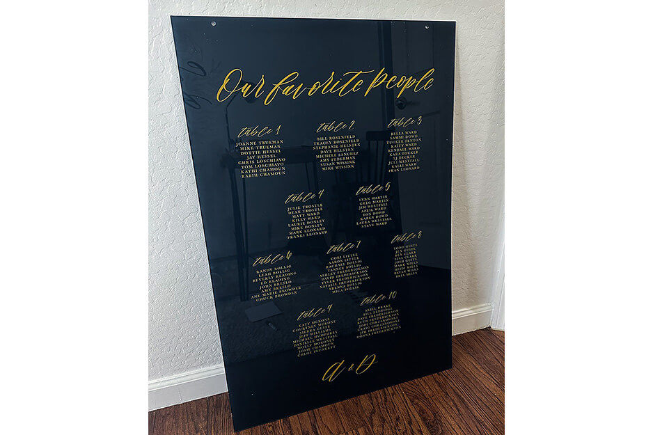 Black acrylic seating chart sign with gold printing leaning against a wall.