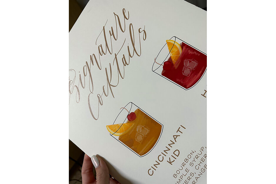 Hand holding white poster with "Signature Cocktails," an image of two drinks and their ingredients printed on it.