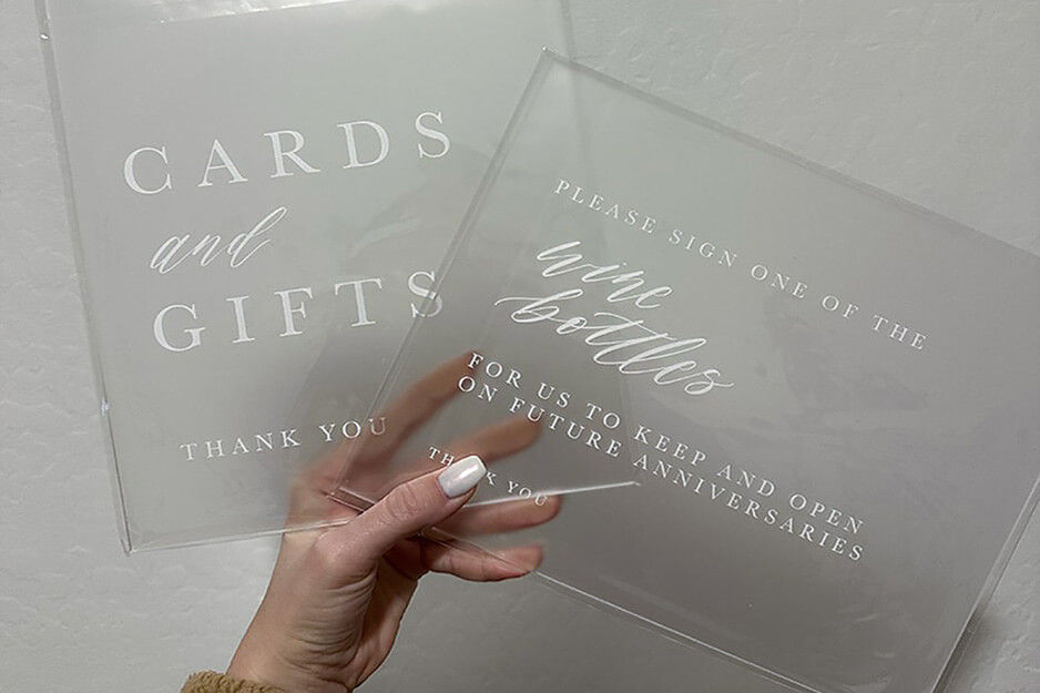 Hand holding two clear acrylic signs with white lettering for "cards and gifts" and "Please sign one of the wine bottles.'