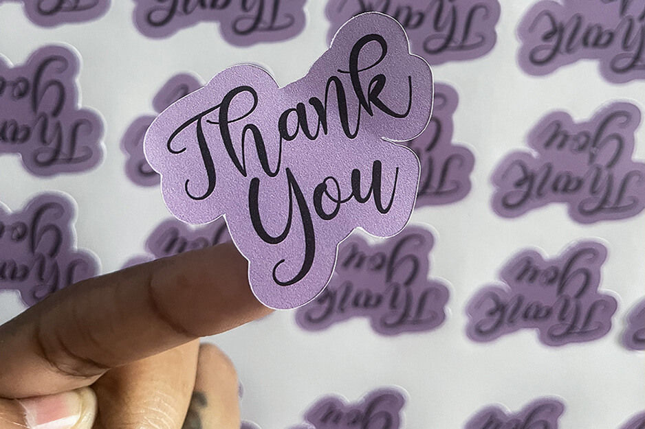Hand holding a "Thank you" sticker in foreground, background is rows of the sticker