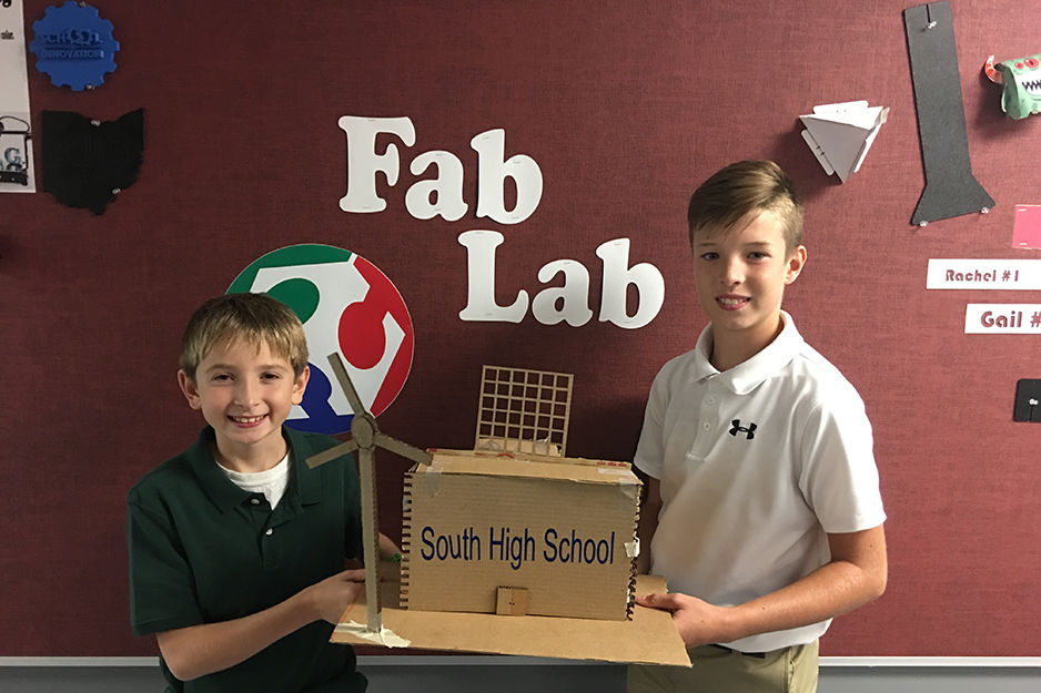 Two students hold a project they built in front of wall graphics for their Fab Lab