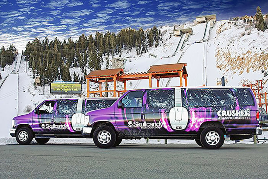 Two vans with purple, black and white Skull Candy graphics, parked in front of a mountain slope.