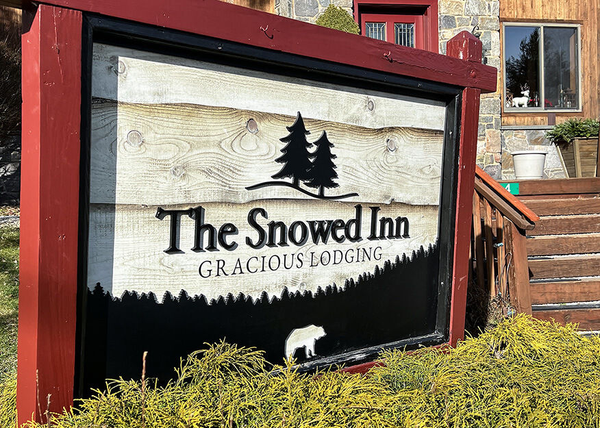 Wooden sign with graphics of pine trees and words "The Snowed Inn"