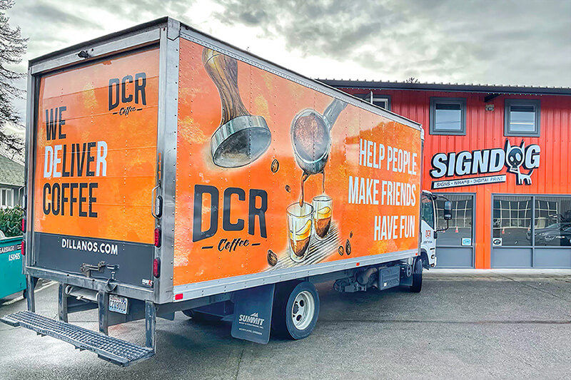 Camión contenedor rotulado en gráficos naranja y negros, estacionado frente al local de SignDog