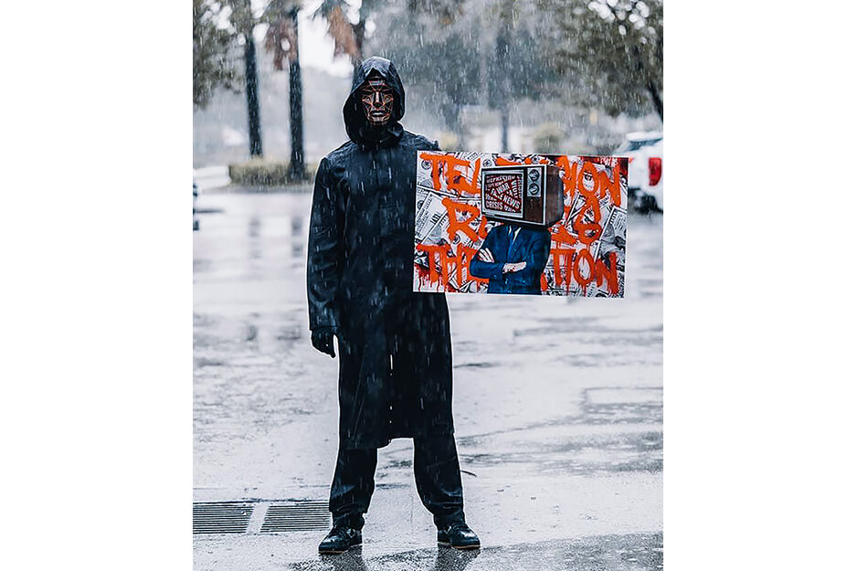 Hombre bajo la lluvia usando una máscara y sujetando un letrero tamaño afiche de colores naranja, rojo y negro.